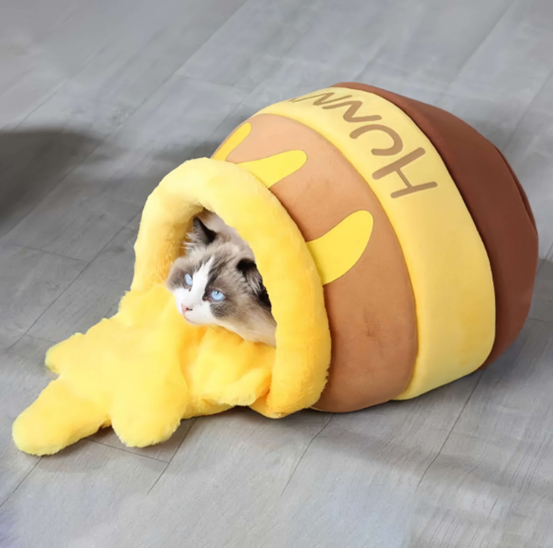 A cat sleeping in a honey jar-shaped cat nest bed in warm yellow and brown tones, featuring a soft plush interior and bee-themed accents.